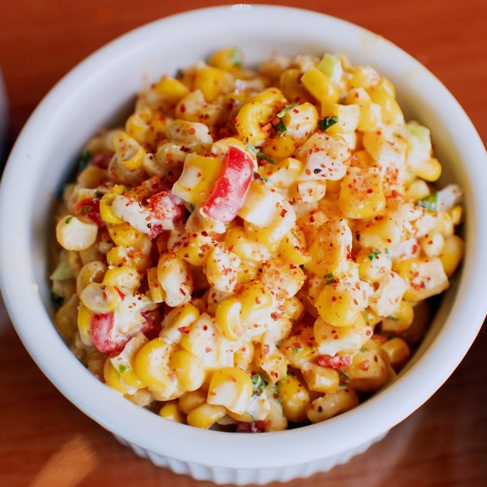 Creamy corn and macaroni salad with red peppers, herbs, and chili seasoning in a white bowl on a wooden table.