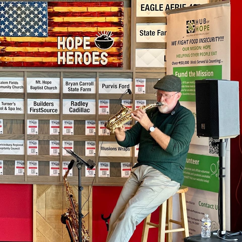 Musician playing saxophone beside a Hope Heroes American flag sign and food insecurity charity banner.