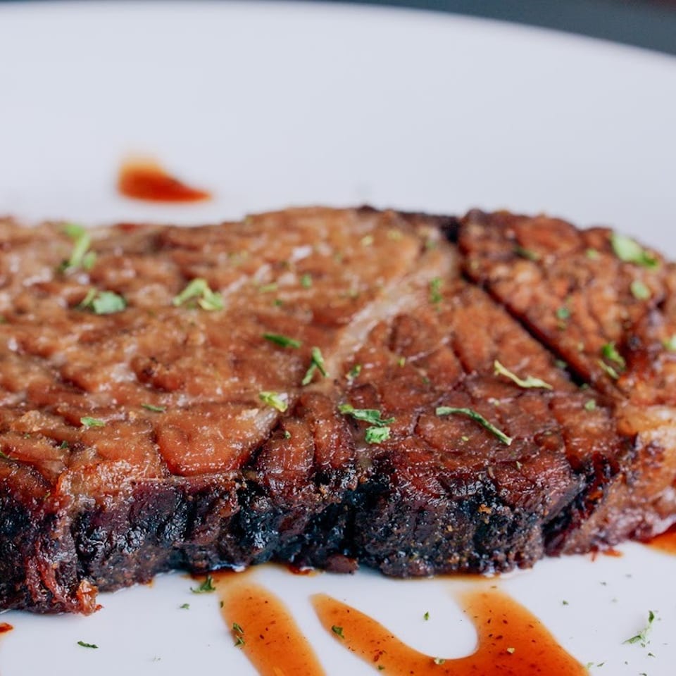 Thick grilled steak topped with chopped herbs and drizzled with reddish sauce on a white plate.