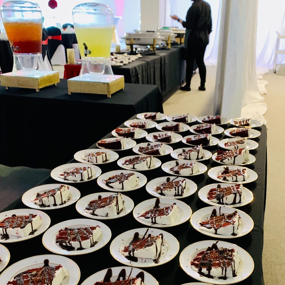 Buffet table displaying numerous cheesecake slices with chocolate drizzle, colorful beverage dispensers, and a server in the background.