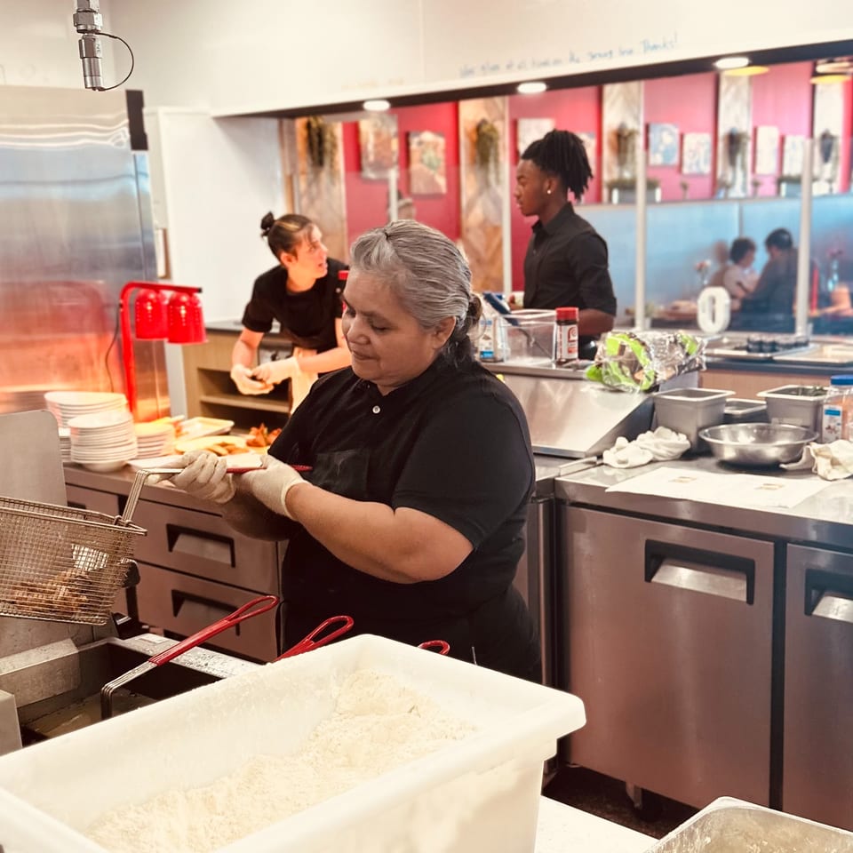 Restaurant kitchen staff prepare food behind the counter, with fryers, ingredients, plates, and colorful dining area visible.