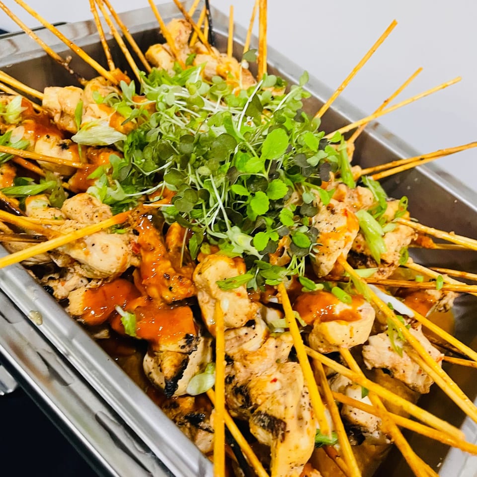 Tray of grilled chicken skewers drizzled with orange sauce and topped with fresh microgreens and scallions.