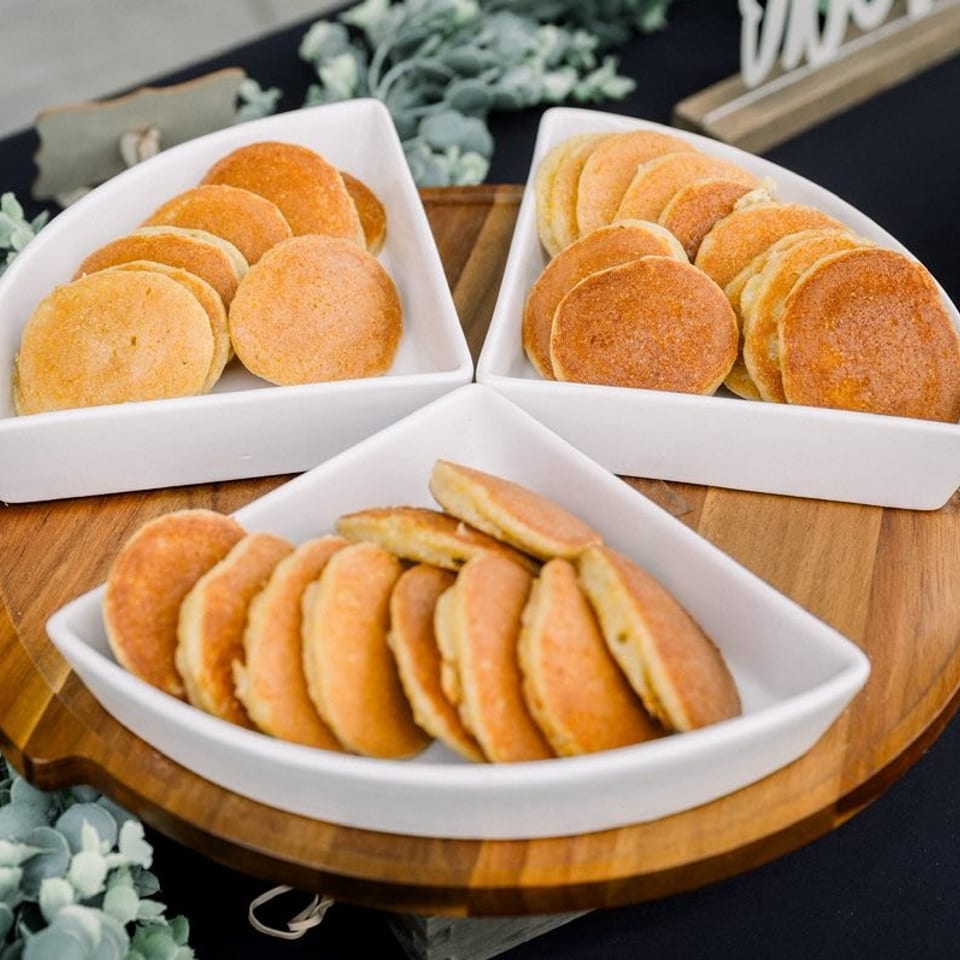 Three white dishes hold stacks of golden-brown round pastries on a wooden serving board surrounded by greenery.