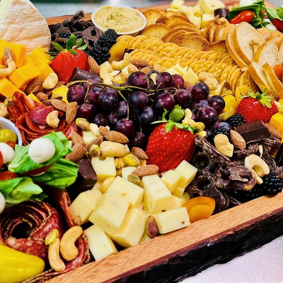 Abundant charcuterie board featuring cheeses, cured meats, crackers, fruit, nuts, olives, vegetables, chocolate, and hummus.