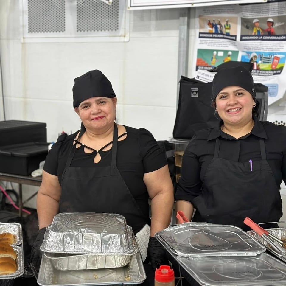 Two smiling food service workers in black uniforms stand behind trays of buns and covered catering dishes.