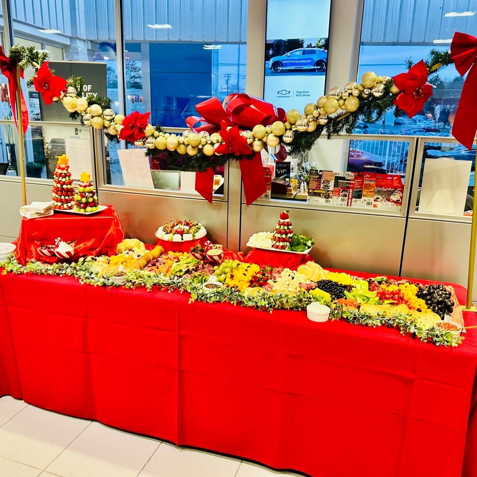 Festive red buffet table displays fruit, vegetables, cheeses, desserts, and chocolate towers beneath gold garlands and red bows.
