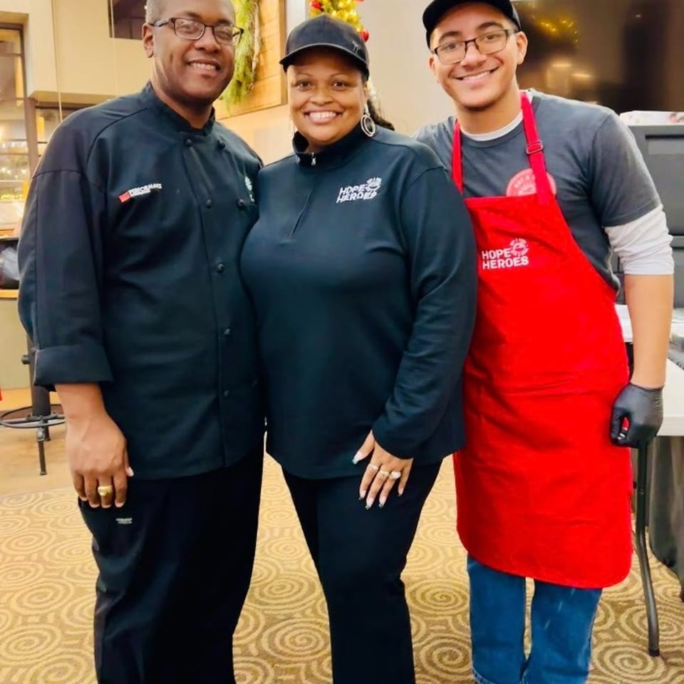 Three smiling restaurant staff members pose together indoors, wearing black uniforms and a red apron beside a decorated Christmas tree.