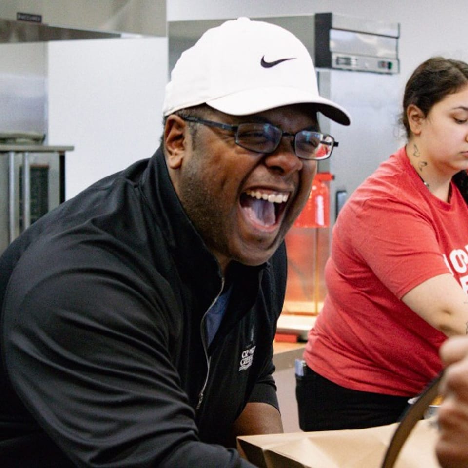 Man in a white Nike cap laughs in a commercial kitchen while a tattooed woman works behind him.
