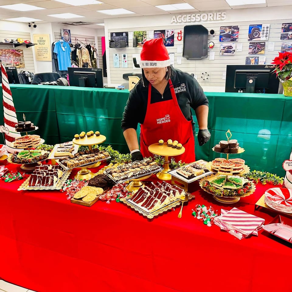 Person in a Santa hat and red apron stands behind a festive table covered with assorted holiday cookies and brownies.