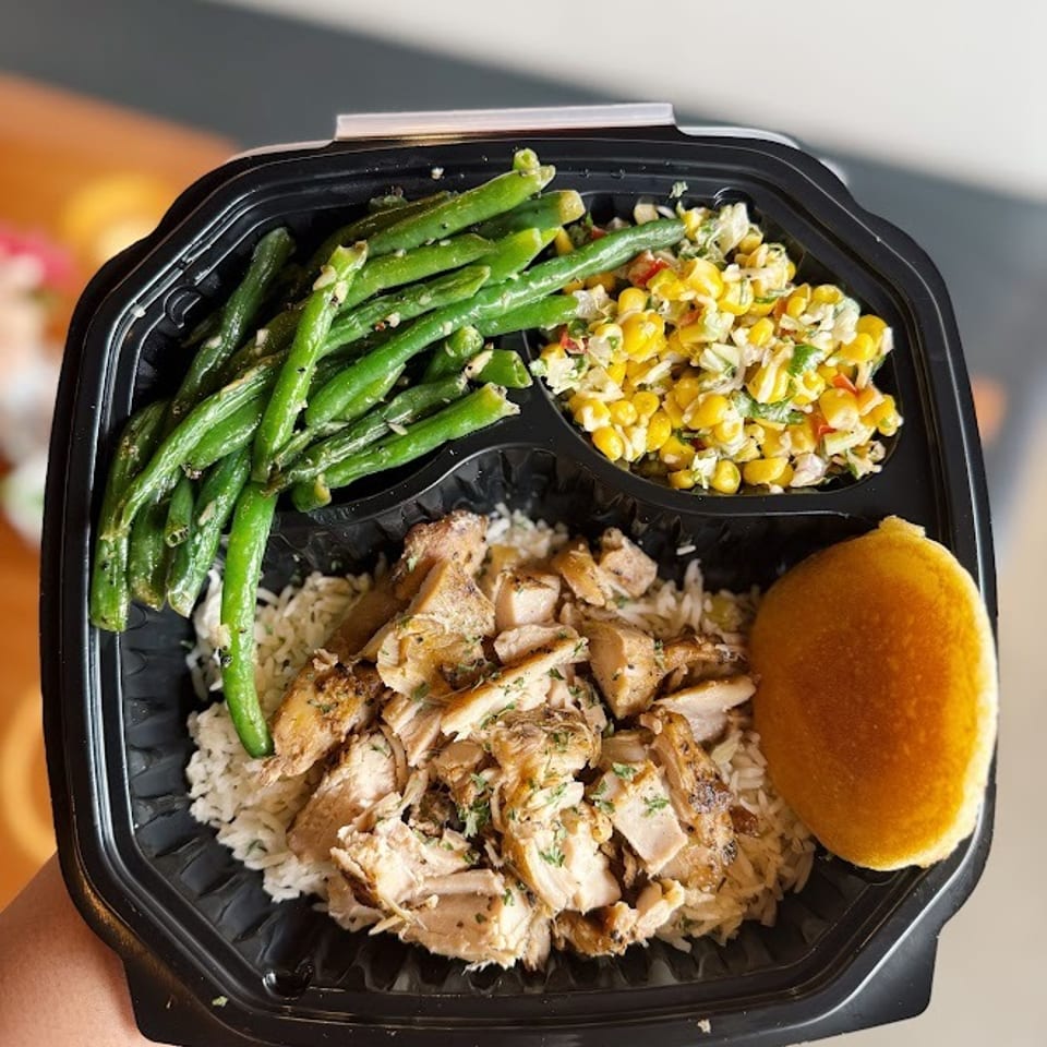Takeout tray with grilled chicken over rice, green beans, corn salad, and a dinner roll.